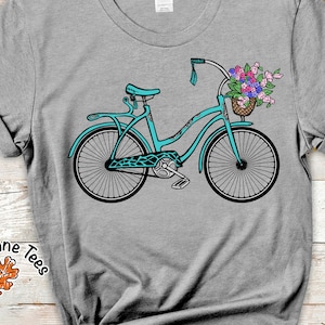 Puede incluir: Una bicicleta azul turquesa con una cesta de flores en el manillar. La bicicleta tiene un asiento blanco y neum&aacute;ticos negros. La bicicleta est&aacute; en una camiseta gris con el texto "Oak Lane Tees" y un dise&ntilde;o de hoja marr&oacute;n.