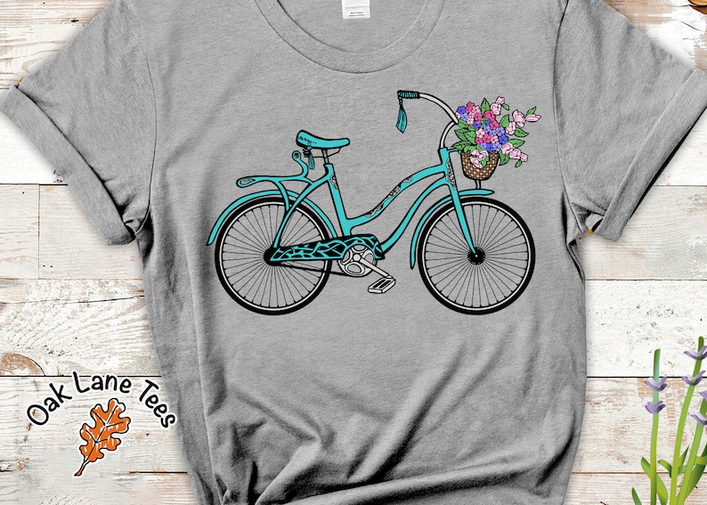 Puede incluir: Una bicicleta azul turquesa con una cesta de flores en el manillar. La bicicleta tiene un asiento blanco y neum&aacute;ticos negros. La bicicleta est&aacute; en una camiseta gris con el texto "Oak Lane Tees" y un dise&ntilde;o de hoja marr&oacute;n.