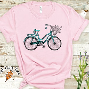 May include: A pink t-shirt with a teal blue bicycle with a basket of flowers on the front. The shirt has a white tag with the word "CANVAS" on it.