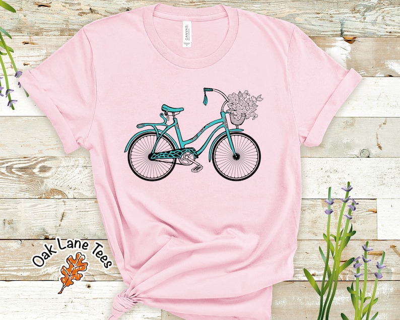May include: A pink t-shirt with a teal blue bicycle with a basket of flowers on the front. The shirt has a white tag with the word "CANVAS" on it.