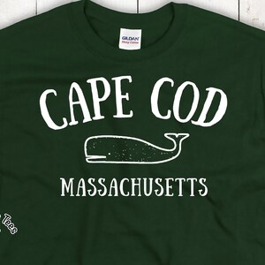 May include: Dark green t-shirt with the words "CAPE COD" in white arched lettering above a white whale illustration. Below the whale is the text "MASSACHUSETTS" in white. The shirt has a Gildan Heavy Cotton tag.
