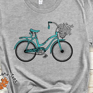 May include: A teal blue bicycle with a basket full of flowers. The bicycle has a white handlebar and a black seat.