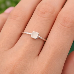 May include: A silver ring with a square, light-colored stone. The ring is simple and elegant, with a thin band and a minimalist design. The square stone is set in a bezel setting, and the ring is shown on a finger.