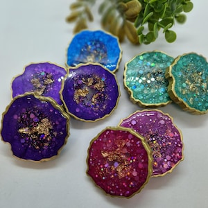 May include: A set of six colourful resin coasters with gold glitter and a gold rim. The coasters are shaped like geodes and have a variety of colours, including purple, blue, green, and pink.