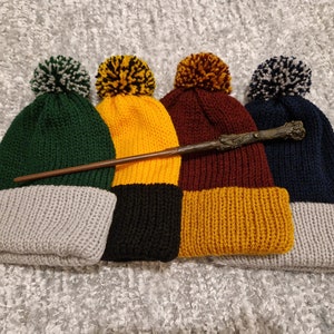 May include: Four knitted hats with pom-poms in green, yellow, maroon, and navy blue. The hats have a contrasting color band at the bottom. A brown wooden wand is lying across the hats.