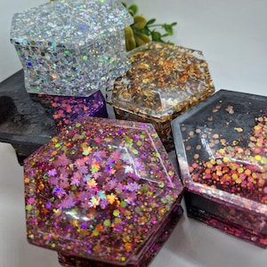 May include: Four hexagonal ring boxes with different decorative finishes. One box is silver with iridescent glitter, one is gold with gold leaf, one is black with iridescent glitter, and one is pink with iridescent glitter.