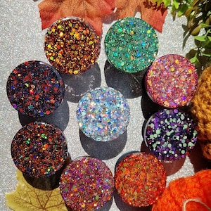 May include: Assortment of ten round phone grips with various glitter patterns. The phone grips are made of clear resin with embedded glitter. The glitter colours include red, orange, green, blue, purple, and iridescent.