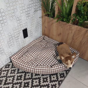 May include: A brown and white checkered dog bed with a plush brown dog toy inside. The bed is in a corner of a room with a patterned wall and a wooden floor.