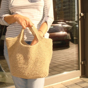 Puede incluir: Un bolso tote beige, tejido a mano, con asas resistentes. El bolso tiene un diseño de tejido abierto y texturizado. El bolso se sostiene frente a una persona que lleva una camisa a rayas y vaqueros. El fondo incluye un edificio y coches aparcados.