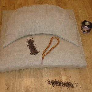 May include: A beige meditation cushion set with a matching bolster, wooden prayer beads, and a small candle holder. Scattered dark seeds add texture. The items are arranged on a wooden floor.