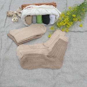 May include: A collection of ribbed socks in various neutral shades including beige, tan, and brown, arranged alongside a woven basket containing rolled socks in beige, gray, green, black, and white, plus a decorative gold-toned bow ornament.