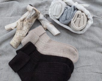 May include: A collection of cozy socks in various colors, including black and beige, are displayed on a gray surface. Rolled socks in white and gray are in a white bag. Decorative birch branches are also present.