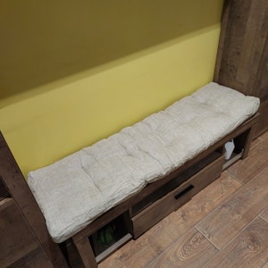 May include: A wooden bench with a light beige, tufted cushion. The bench has a drawer and an open storage space below. The bench is approximately 50 inches long and is placed against a yellow wall.