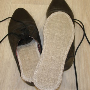 Può includere: Scarpe in pelle nera con lacci neri, una con una soletta interna rimovibile in lino beige. La soletta è visibile in una scarpa e separata dall'altra. Le scarpe sono su una superficie di legno chiaro.