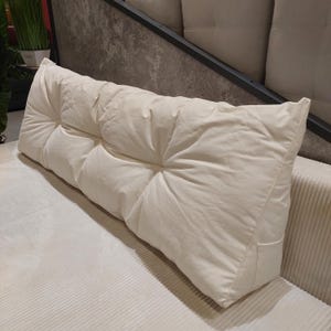 May include: A long, rectangular, off-white wedge pillow featuring a tufted design with multiple recessed buttons across its surface. The pillow has a tapered shape, a smooth fabric texture, and includes a small side pocket on the right end.