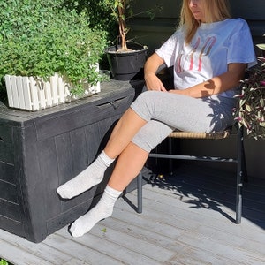 May include: A woman wearing a white t-shirt with the word "GAP" printed on it, gray capri pants, and white socks. She is sitting on a chair with a wooden seat and metal legs. The chair is in front of a black storage box with a white picket fence design.
