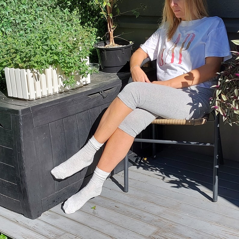 May include: A woman wearing a white t-shirt with the word "GAP" printed on it, gray capri pants, and white socks. She is sitting on a chair with a wooden seat and metal legs. The chair is in front of a black storage box with a white picket fence design.