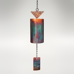 May include: A handcrafted copper rain chain with a cylindrical body and a triangular top piece. The body features a blend of green, red, and blue hues. A rectangular piece hangs at the bottom. The chain is suspended from a black chain.