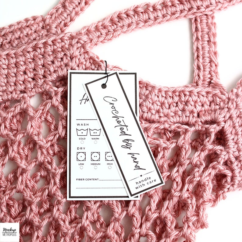 K&ouml;nnte beinhalten: Ein wei&szlig;es Etikett mit schwarzem Text und Symbolen f&uuml;r Waschanweisungen. Das Etikett lautet "Crocheted by hand, handle with care". Das Etikett ist an einem rosafarbenen H&auml;kelartikel befestigt.