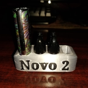 May include: A white 3D printed stand with the word "Novo 2" printed on the front. The stand has three slots for holding small cylindrical objects. Two black cylindrical objects are in the slots. A third slot is empty. A multi-colored cylindrical object is standing upright behind the stand.