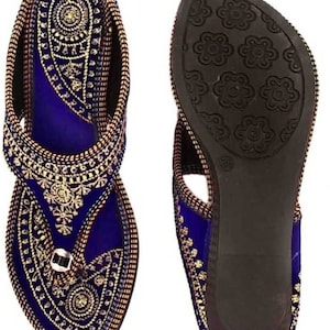 May include: A pair of blue and gold embroidered sandals. The sandals feature intricate gold threadwork on a blue velvet base. The soles are dark brown with a floral pattern. The sandals are designed with a toe loop and ankle strap.