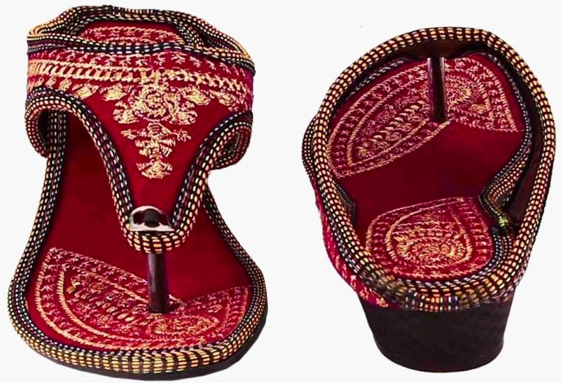 May include: Pair of red and gold embroidered sandals. The sandals feature intricate gold embroidery on a red background, with a black and gold braided trim. The design includes a toe post and a small heel.