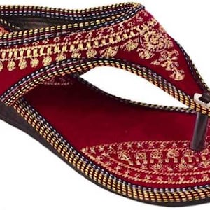 May include: Burgundy wedge sandal with intricate gold embroidery. The sandal features a thong-style design with a dark brown sole and a decorative pattern around the edges. A stylish footwear option.