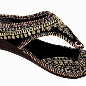 May include: Black wedge sandal with intricate gold embroidery. The sandal features a thong strap design and a brown wedge heel. The upper is adorned with detailed patterns and embellishments, creating a decorative and stylish footwear option.