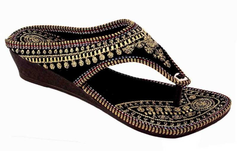 May include: Black wedge sandal with intricate gold embroidery. The sandal features a thong strap design and a brown wedge heel. The upper is adorned with detailed patterns and embellishments, creating a decorative and stylish footwear option.