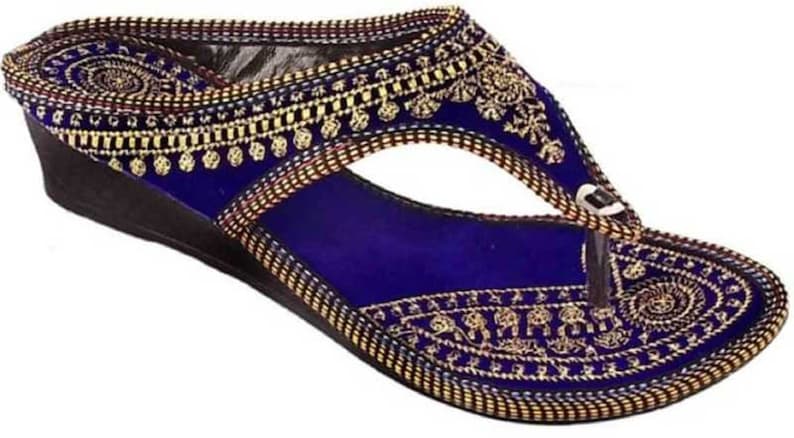 May include: A blue sandal with a wedge heel. The sandal features intricate gold embroidery along the edges and straps, with a decorative pattern on the footbed. The thong strap is white and the sole is black.