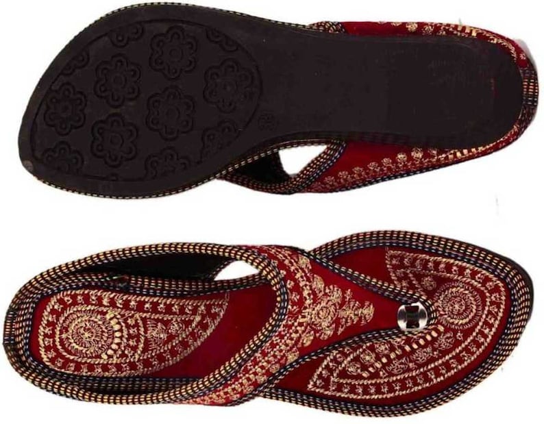 May include: Burgundy and gold embroidered sandals. The sandals feature intricate gold threadwork on a burgundy base, with a black sole. The design includes a toe loop and a decorative button. The sole has a floral pattern.