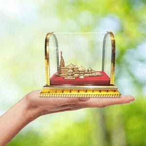May include: A gold-coloured miniature temple model inside a clear glass display case with a red velvet base. The display case has gold trim and is held in a hand against a blurred green background.