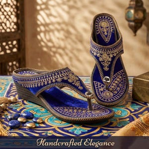 May include: A pair of blue velvet sandals with gold embroidery and a wedge heel. The sandals feature intricate patterns and are displayed on a patterned table with decorative elements. The image includes the text "Handcrafted Elegance".