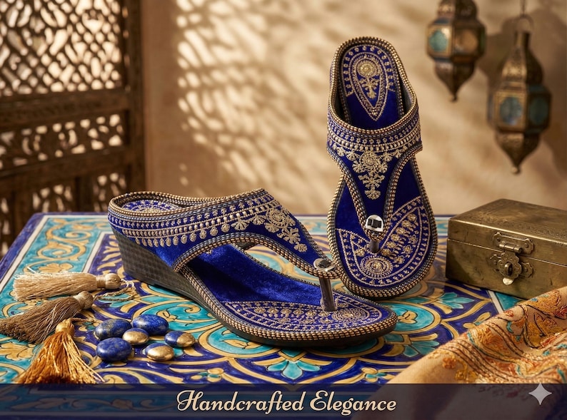 May include: A pair of blue velvet sandals with gold embroidery and a wedge heel. The sandals feature intricate patterns and are displayed on a patterned table with decorative elements. The image includes the text "Handcrafted Elegance".