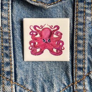 May include: A pink octopus with eight tentacles and large eyes is illustrated on a white square pin.
