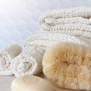 May include: A stack of white textured cotton washcloths, a round beige natural fiber scrubbing brush, and a smooth oval-shaped bar of soap. The washcloths feature a waffle-like weave pattern, while the brush has dense, light brown bristles.