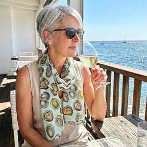 May include: A light blue scarf with an oyster print is draped around the neck of a person wearing sunglasses. The person is holding a glass of white wine, with the ocean in the background. The scarf features a repeating pattern of oyster illustrations.