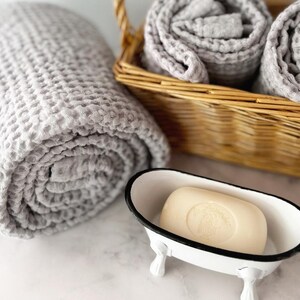 May include: A light gray waffle-textured towel rolled next to a woven wicker basket containing two additional rolled gray towels and a cream-colored bar of soap resting inside a small white metal bathtub-shaped dish with black trim.