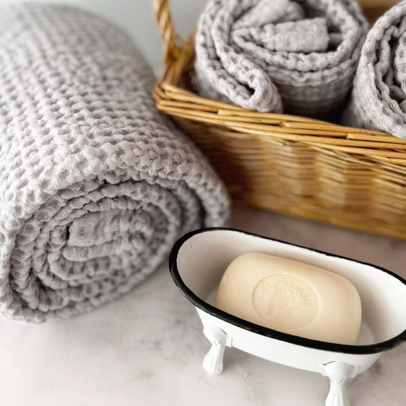 May include: A light gray waffle-textured towel rolled next to a woven wicker basket containing two additional rolled gray towels and a cream-colored bar of soap resting inside a small white metal bathtub-shaped dish with black trim.