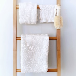 May include: A light brown wooden ladder towel rack holding two white textured towels and a smaller white ruffled cloth. A round beige bath brush with a long wooden handle hangs from the top rung next to the smaller cloth.