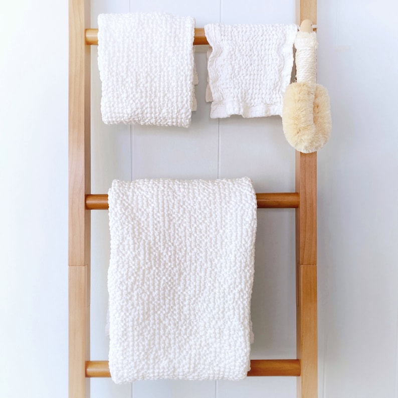 May include: A light brown wooden ladder towel rack holding two white textured towels and a smaller white ruffled cloth. A round beige bath brush with a long wooden handle hangs from the top rung next to the smaller cloth.