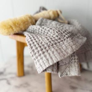 May include: A light gray waffle-weave towel with a textured grid pattern draped over a wooden stool, accompanied by a long-handled beige bath brush with natural bristles and a looped cord for hanging, creating a neutral-toned bathroom set.