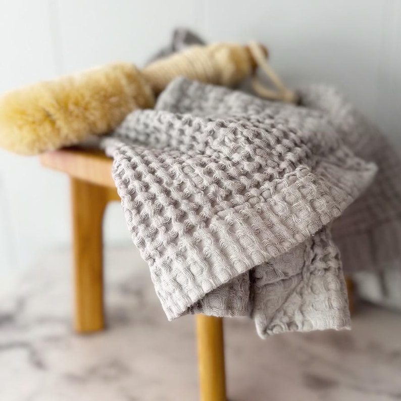 May include: A light gray waffle-weave towel with a textured grid pattern draped over a wooden stool, accompanied by a long-handled beige bath brush with natural bristles and a looped cord for hanging, creating a neutral-toned bathroom set.