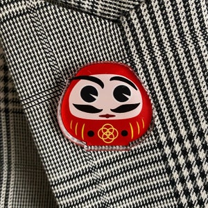 May include: A red and yellow daruma doll pin with a white face, black eyes, and a black mustache. The daruma doll is a traditional Japanese good luck charm.