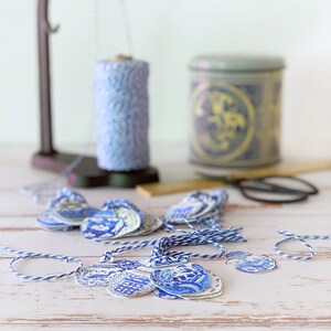 Puede incluir: Una colecci&oacute;n de etiquetas de regalo con estampado azul y blanco con cuerda atada. Las etiquetas est&aacute;n dispuestas sobre una superficie de madera blanca con un carrete de cuerda y un recipiente de cer&aacute;mica azul y blanco en el fondo.