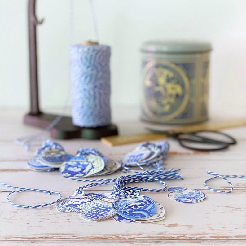 Puede incluir: Una colecci&oacute;n de etiquetas de regalo con estampado azul y blanco con cuerda atada. Las etiquetas est&aacute;n dispuestas sobre una superficie de madera blanca con un carrete de cuerda y un recipiente de cer&aacute;mica azul y blanco en el fondo.
