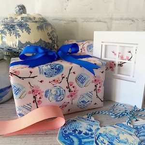 May include: A pink gift box wrapped in blue and white patterned paper with a blue ribbon. The paper features a design of blue and white porcelain jars with pink cherry blossoms. The box is sitting on a white surface with two blue and white porcelain jar gift tags.