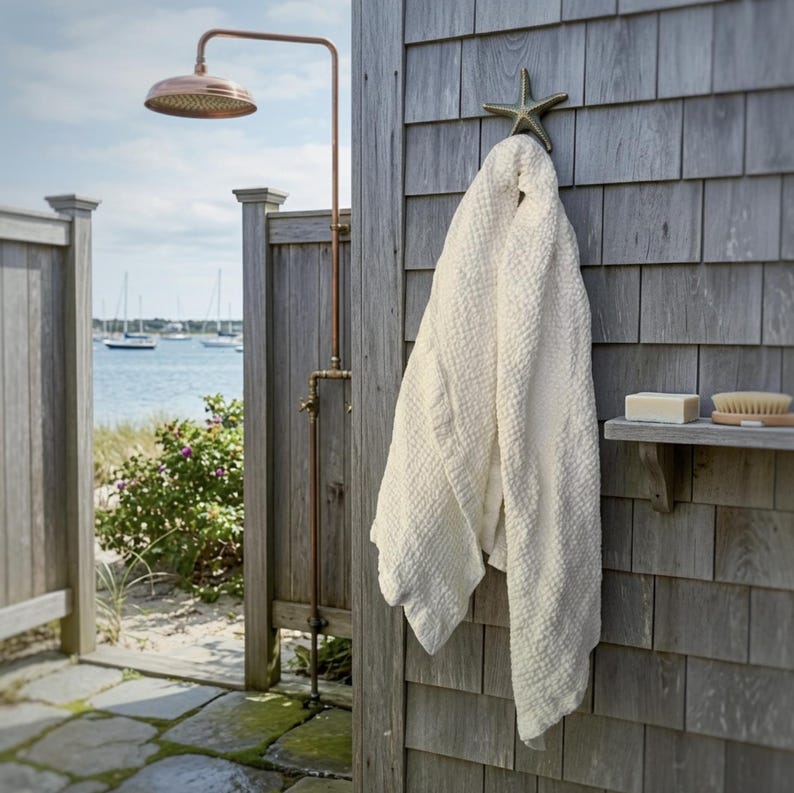May include: A white waffle-weave towel hangs from a bronze-colored starfish hook mounted on a gray shingled wall. Nearby, a copper outdoor shower fixture stands tall, and a small wooden shelf holds a bar of soap and a wooden scrub brush.
