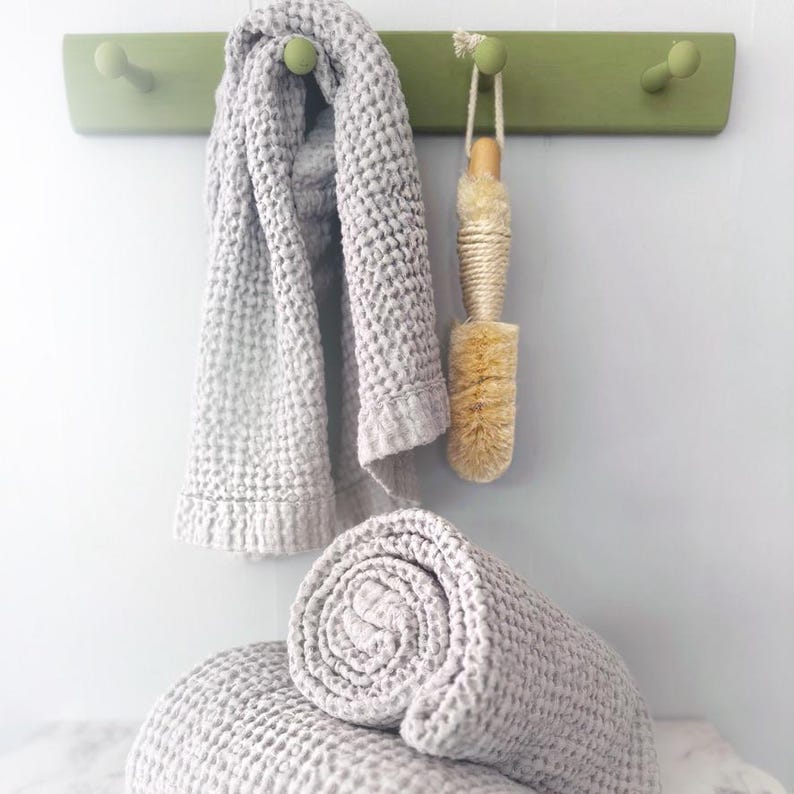 May include: A light gray waffle-weave towel hanging on a green wooden peg rack next to a natural fiber bath brush with a rope loop. Below, a second matching light gray waffle-weave towel is rolled up, showcasing the textured fabric pattern.