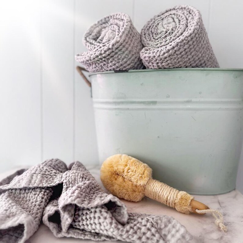 May include: Two rolled light gray waffle-weave towels sit inside a pale mint green metal bucket. A third matching waffle-weave towel is draped in front of the bucket next to a round wooden bath brush with natural tan bristles and a rope handle.